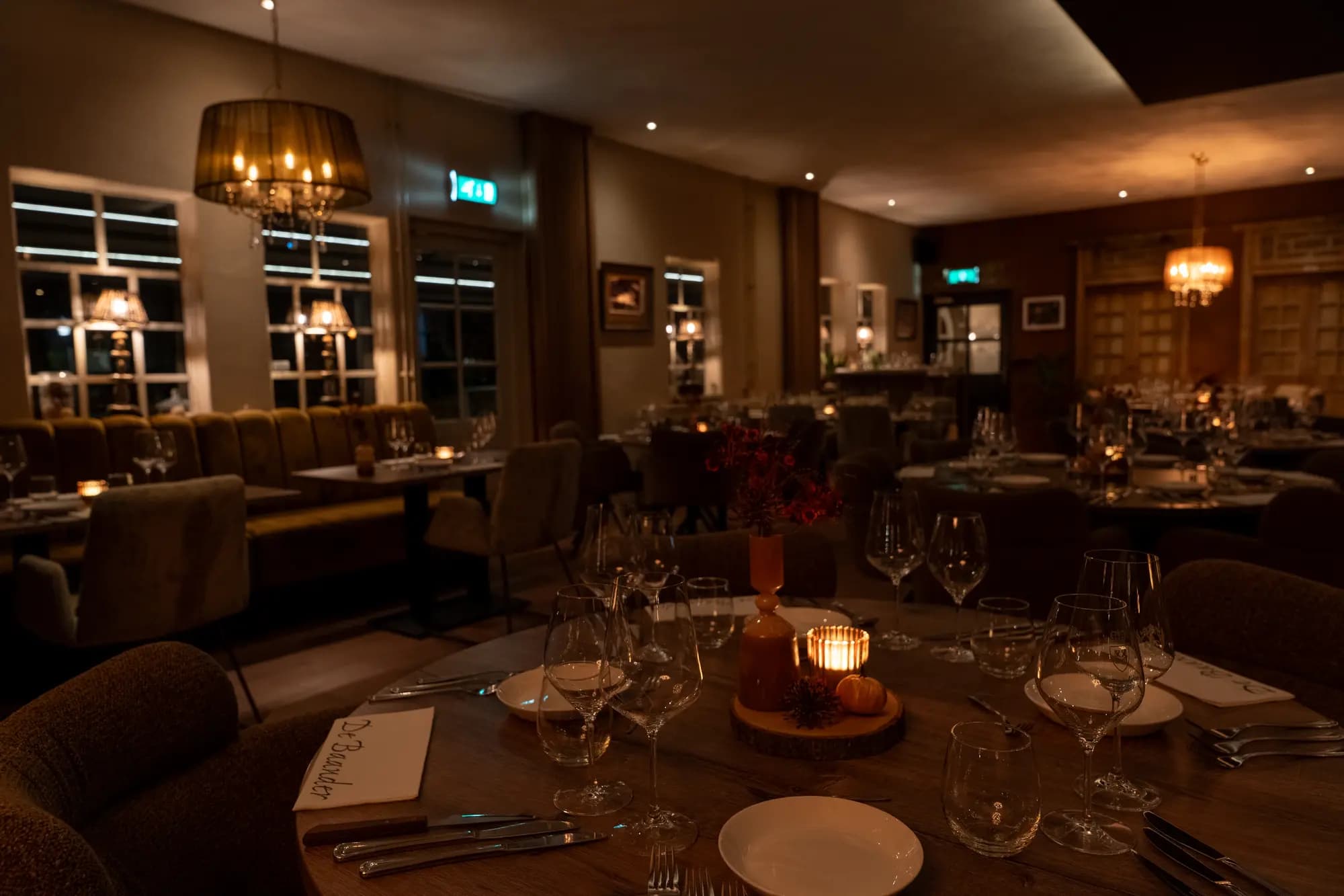 Gedekte ronde tafel met wijnglazen, kaars en droogbloemen, verlichte eetzaal op de achtergrond