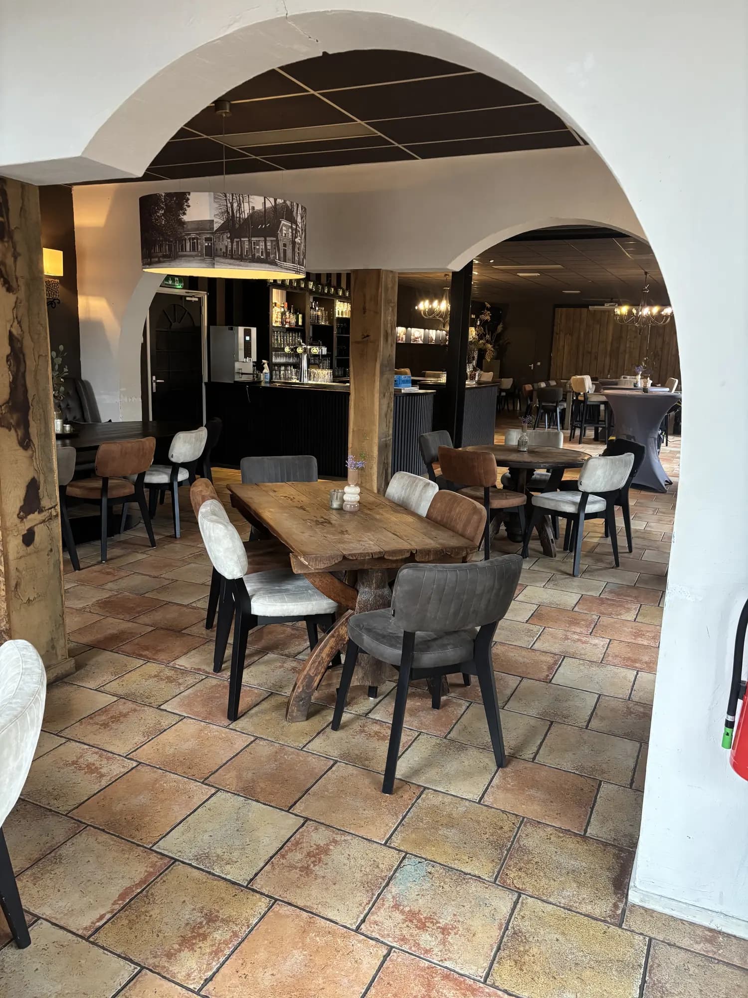 Doorkijk door een gewelfboog naar de bar, met houten eettafel en stoelen op de voorgrond