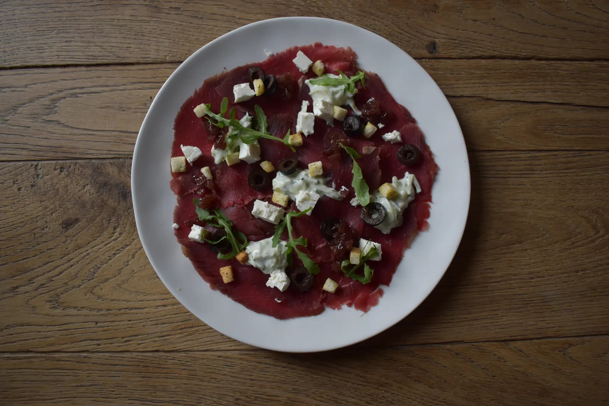 Rundercarpaccio met feta, rucola, zwarte olijven en croutons op een wit bord