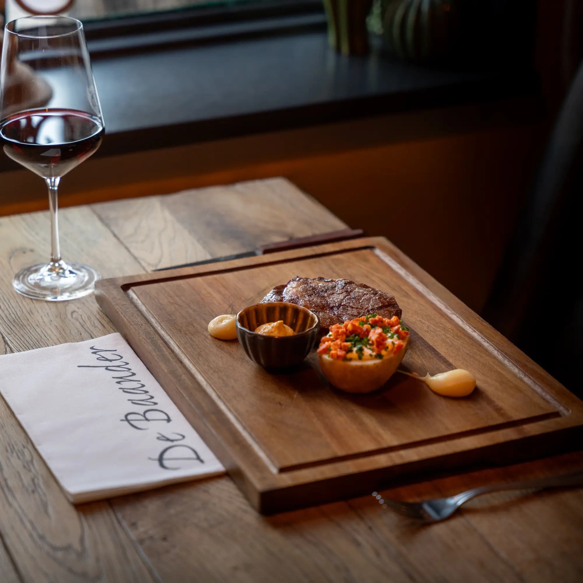 Gegrilde dry-aged steak op houten plank met sausjes, naast een glas rode wijn