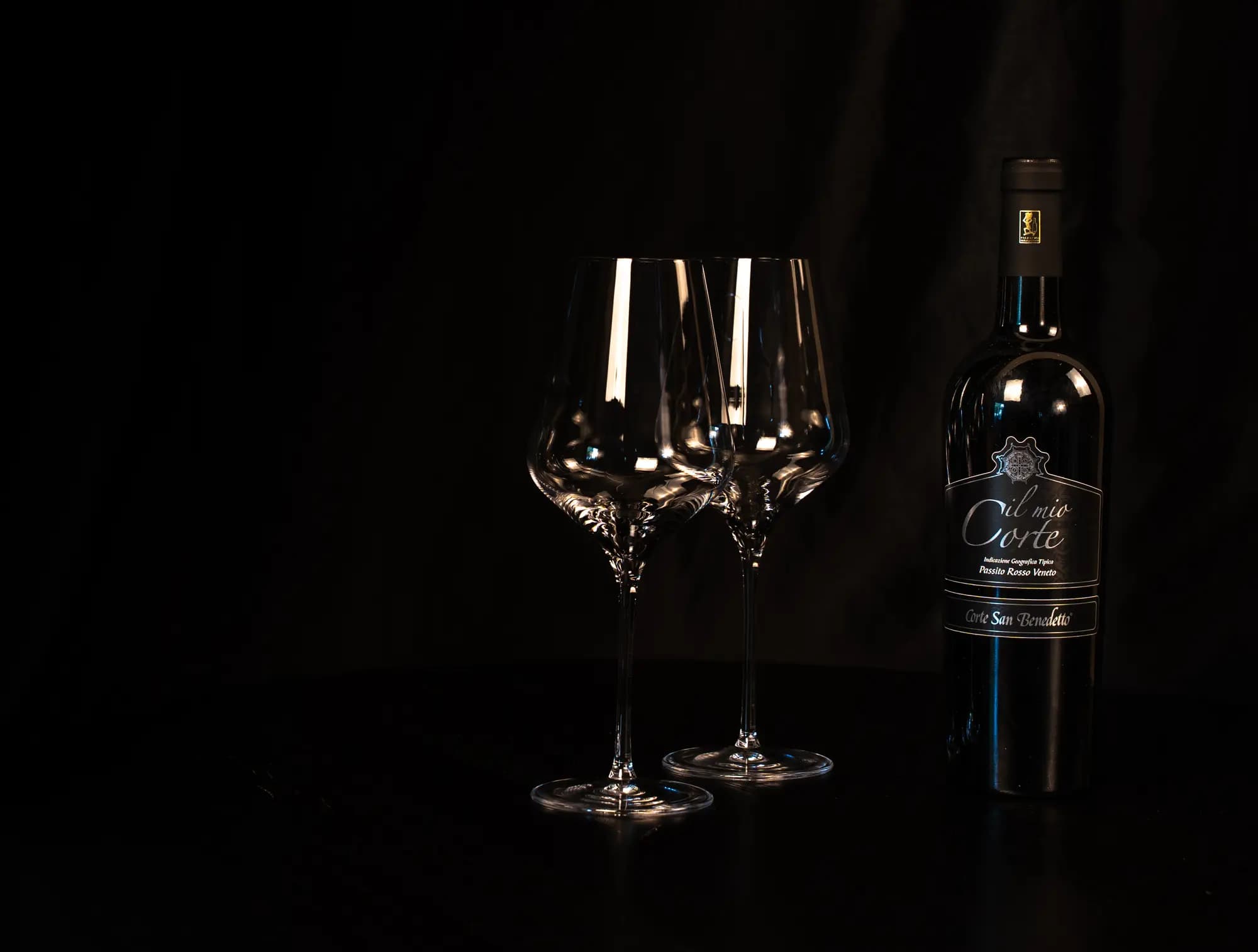 Fles Il Mio Corte Passito Rosso naast twee lege wijnglazen op een donkere tafel