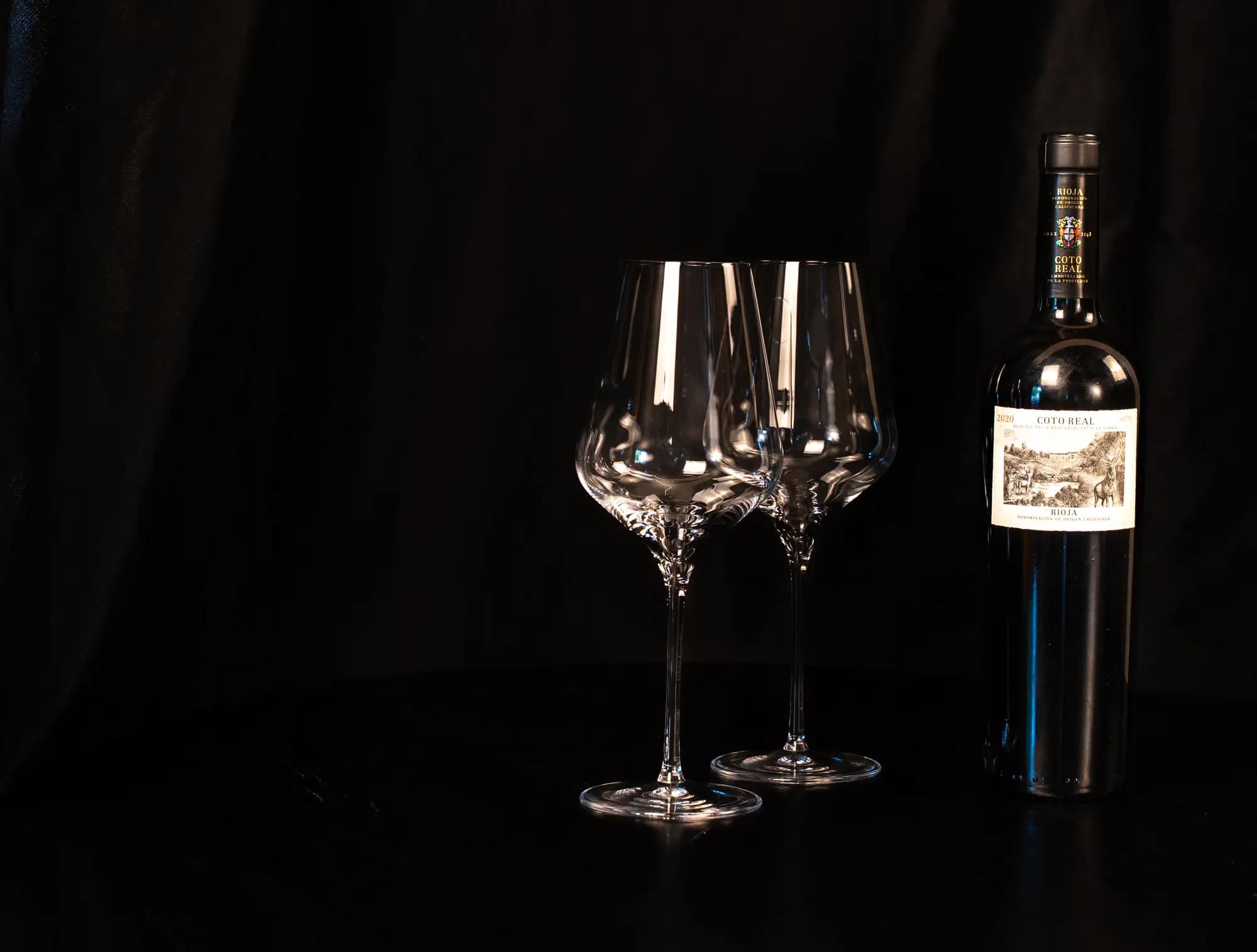 Fles Coto Real Rioja naast twee lege wijnglazen op een donkere tafel