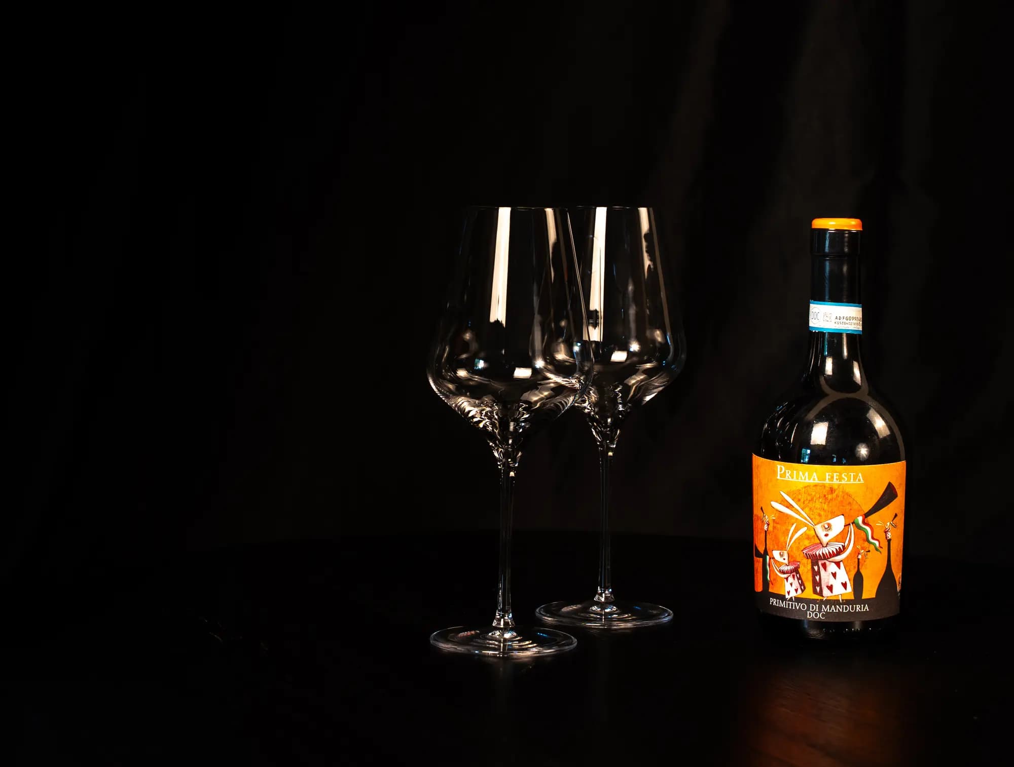 Fles Prima Festa Primitivo di Manduria naast twee lege wijnglazen op een donkere tafel