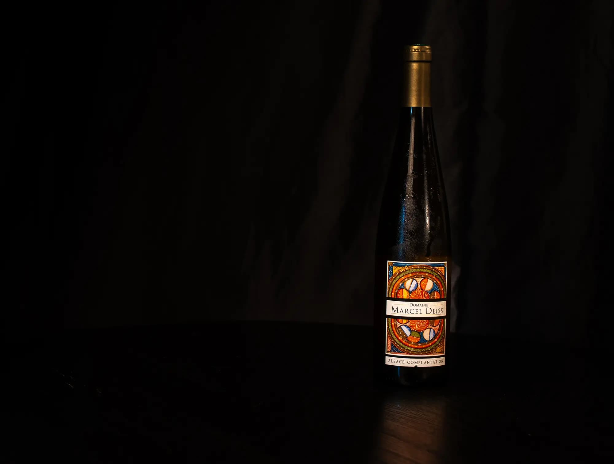 Fles Domaine Marcel Deiss Alsace Complantation met kleurrijk mozaiek-etiket tegen zwarte achtergrond