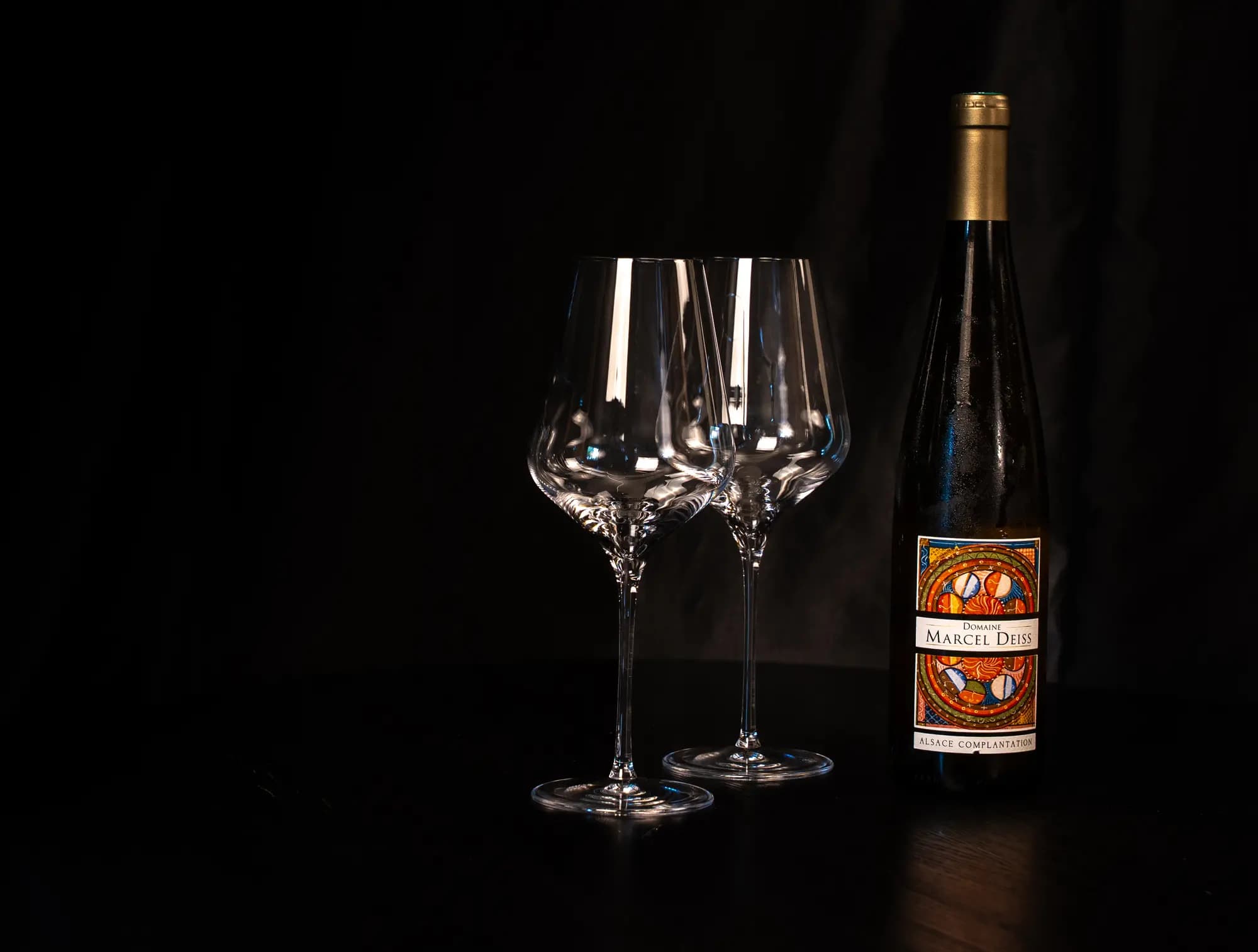 Fles Marcel Deiss Alsace Complantation naast twee lege wijnglazen op een donkere tafel