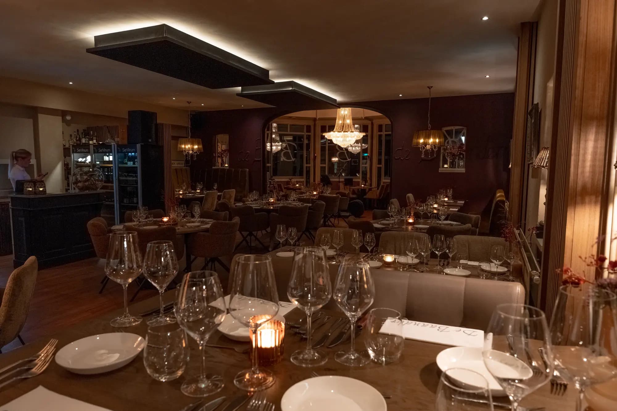 Overzicht van de eetzaal met verlichte bar, gedekte tafels en kaarsjes in warm avondlicht